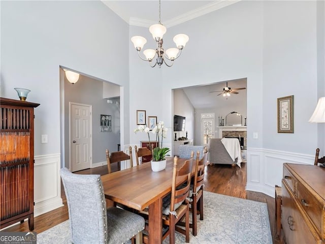 dining area featuring a stone fireplace, a decorative wall, ceiling fan with notable chandelier, wood finished floors, and crown molding