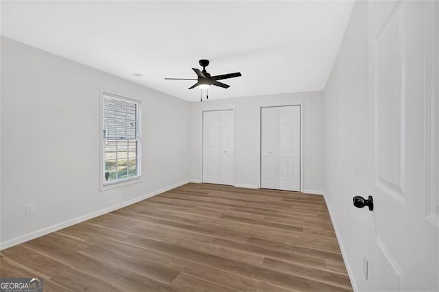 unfurnished bedroom featuring two closets, ceiling fan, baseboards, and wood finished floors