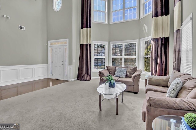 carpeted living room featuring a wainscoted wall, visible vents, a towering ceiling, and a decorative wall