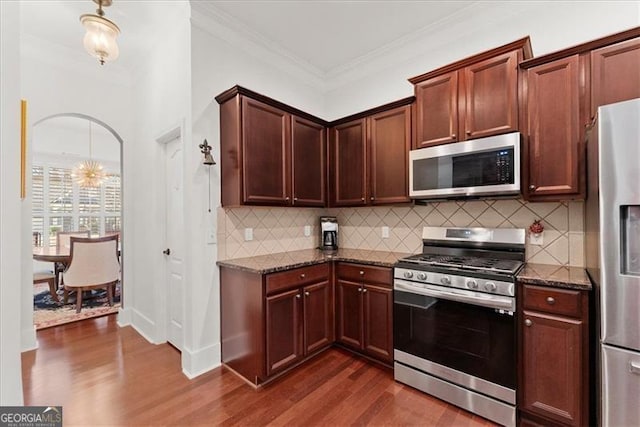 kitchen with arched walkways, dark wood-type flooring, appliances with stainless steel finishes, ornamental molding, and dark stone countertops