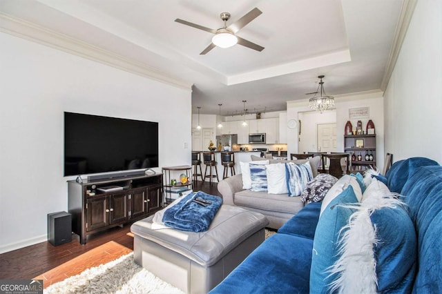 living area featuring ceiling fan with notable chandelier, baseboards, a tray ceiling, dark wood finished floors, and crown molding