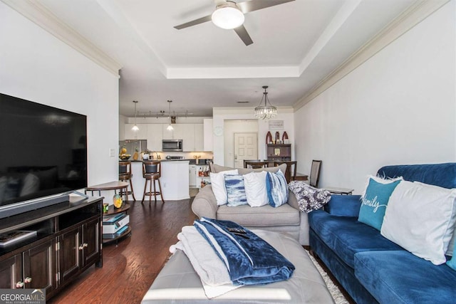 living area featuring dark wood-type flooring, a tray ceiling, ornamental molding, and ceiling fan with notable chandelier