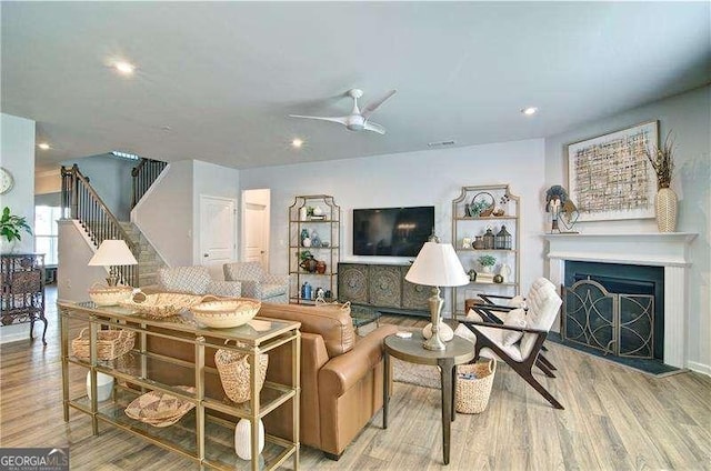 living room featuring stairs, a fireplace with raised hearth, wood finished floors, and a ceiling fan