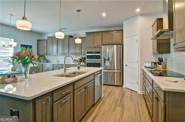 kitchen with stainless steel appliances, light wood-style floors, a kitchen island with sink, a sink, and wall chimney range hood