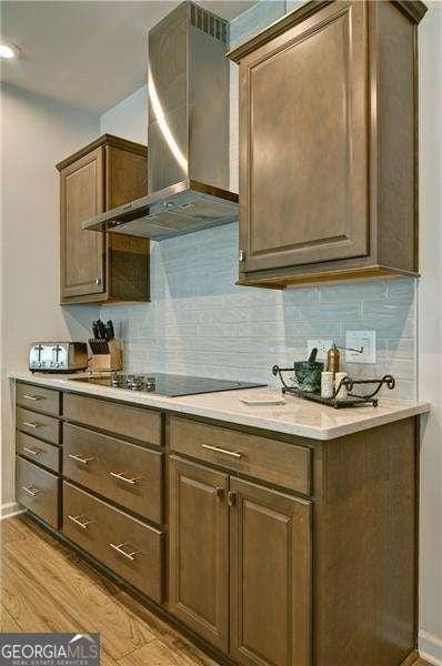 kitchen featuring black electric stovetop, light countertops, wall chimney range hood, light wood-type flooring, and decorative backsplash