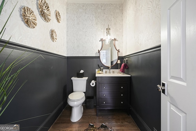 half bathroom featuring a wainscoted wall, toilet, vanity, wood finished floors, and wallpapered walls