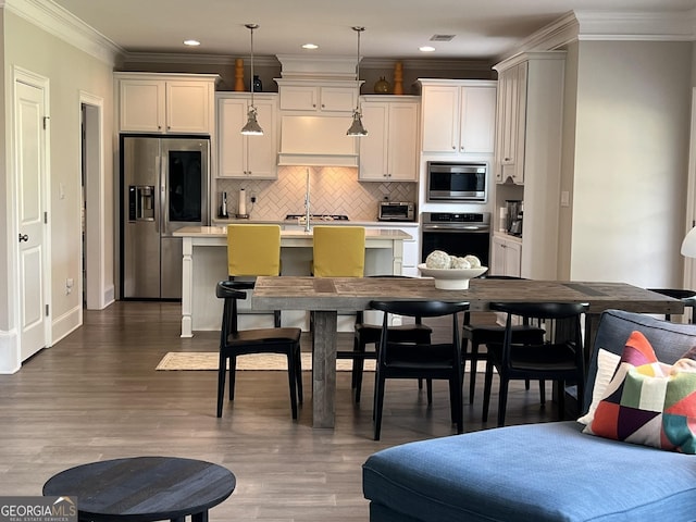 kitchen with crown molding, custom exhaust hood, stainless steel appliances, light countertops, and an island with sink