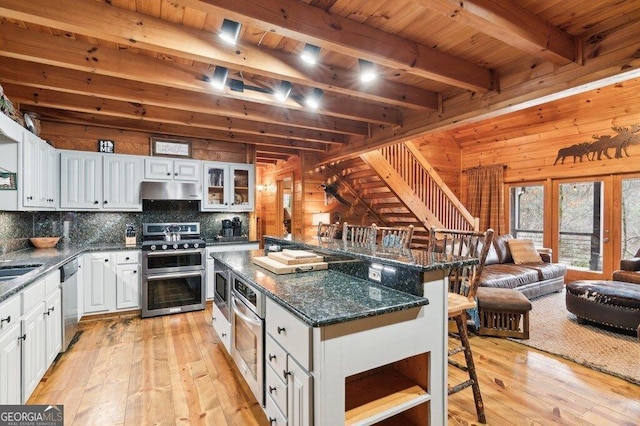 kitchen featuring beam ceiling, appliances with stainless steel finishes, wooden walls, light wood-type flooring, and a kitchen bar