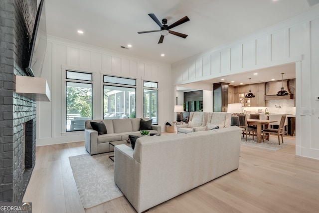 living area with a decorative wall, a ceiling fan, visible vents, a brick fireplace, and light wood-type flooring