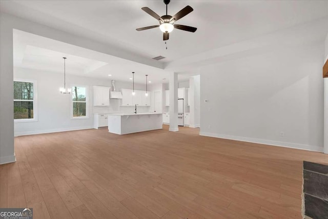 unfurnished living room with light wood-type flooring, baseboards, a tray ceiling, and ceiling fan with notable chandelier