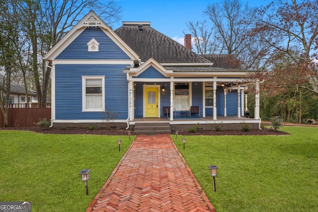 victorian-style house featuring a chimney, a porch, a shingled roof, a front yard, and fence