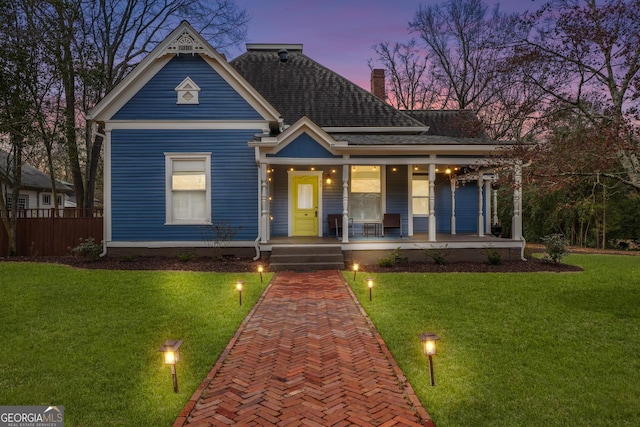 victorian-style house featuring a porch, a shingled roof, fence, a front lawn, and a chimney