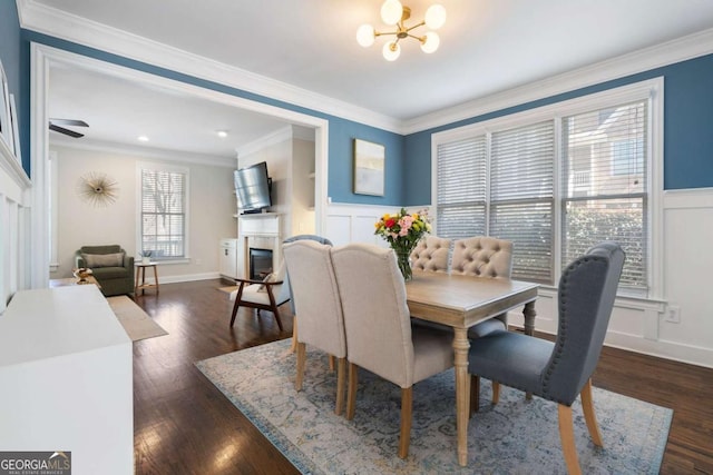 dining area featuring a wainscoted wall, dark wood finished floors, a notable chandelier, ornamental molding, and a glass covered fireplace