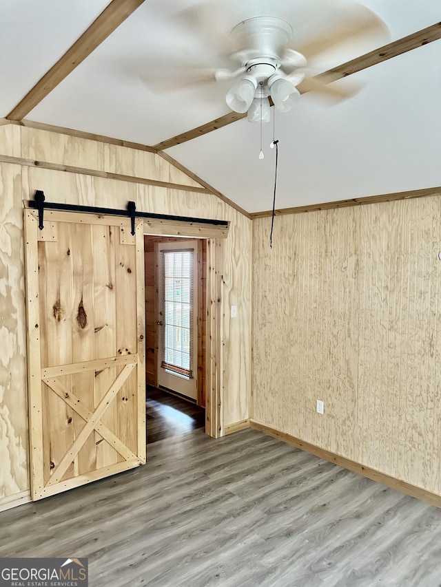 spare room featuring lofted ceiling with beams, a barn door, wooden walls, wood finished floors, and a ceiling fan