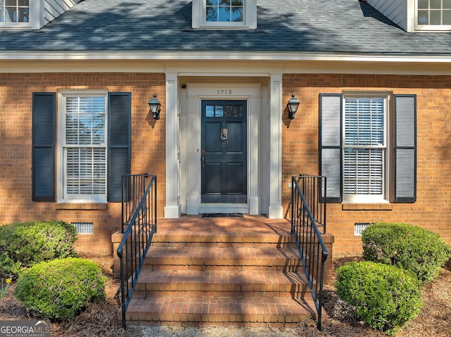 view of exterior entry featuring roof with shingles, brick siding, and crawl space