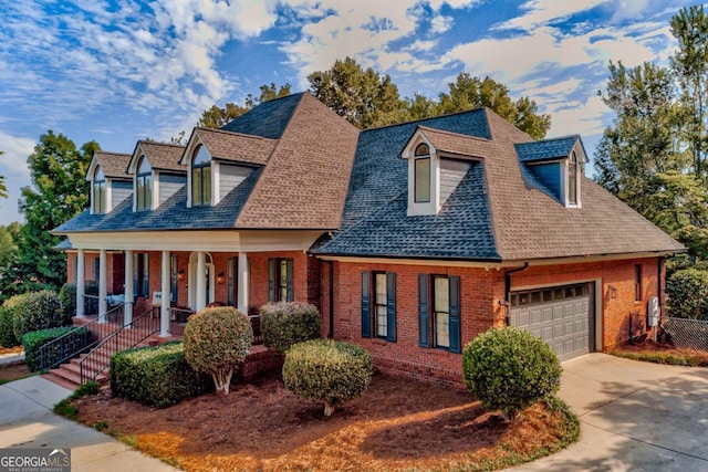 new england style home with driveway, a shingled roof, covered porch, a garage, and brick siding