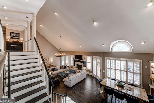 living room featuring a stone fireplace, a notable chandelier, stairs, dark wood finished floors, and decorative columns