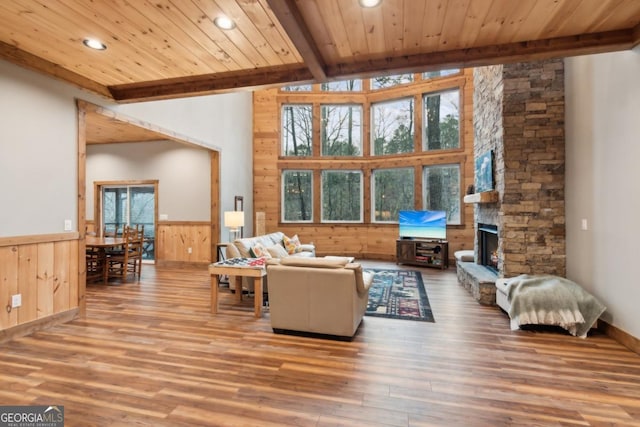 living room featuring a stone fireplace, beamed ceiling, wainscoting, and wood ceiling