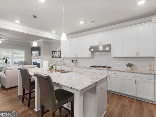 kitchen with white cabinets, open floor plan, under cabinet range hood, stainless steel gas stovetop, and a sink