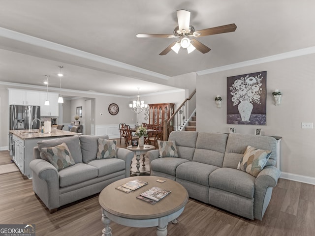 living room featuring ceiling fan with notable chandelier, wood finished floors, baseboards, stairway, and crown molding