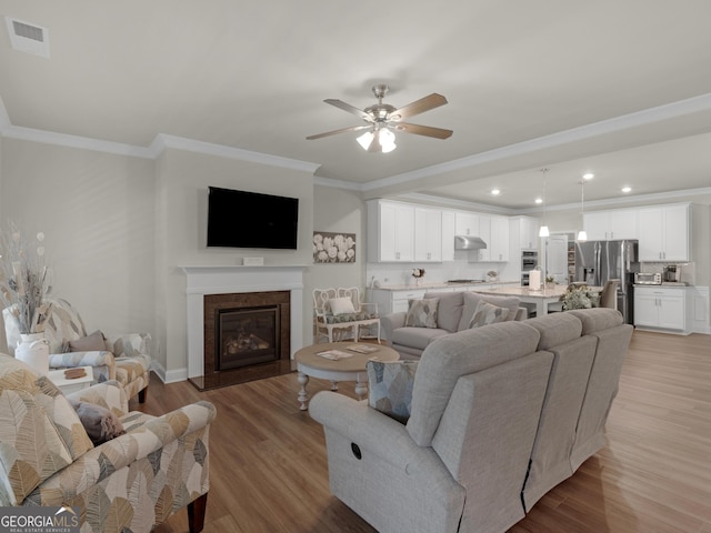 living room featuring visible vents, a ceiling fan, a glass covered fireplace, ornamental molding, and light wood-style floors