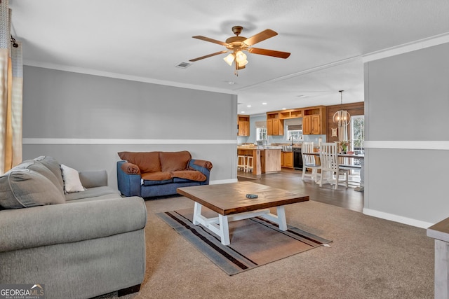 carpeted living room with ceiling fan with notable chandelier, visible vents, baseboards, and ornamental molding