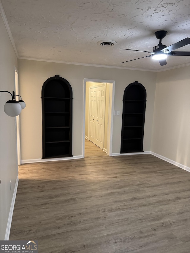 unfurnished room with a textured ceiling, built in shelves, wood finished floors, visible vents, and crown molding