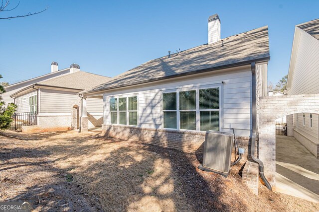 rear view of house with brick siding, a shingled roof, fence, central AC, and a chimney