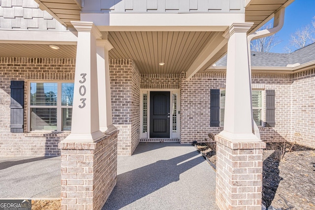 entrance to property featuring covered porch, a shingled roof, and brick siding