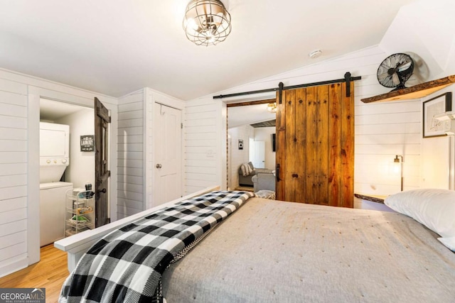 bedroom featuring a barn door, lofted ceiling, wood walls, light wood-type flooring, and stacked washing maching and dryer