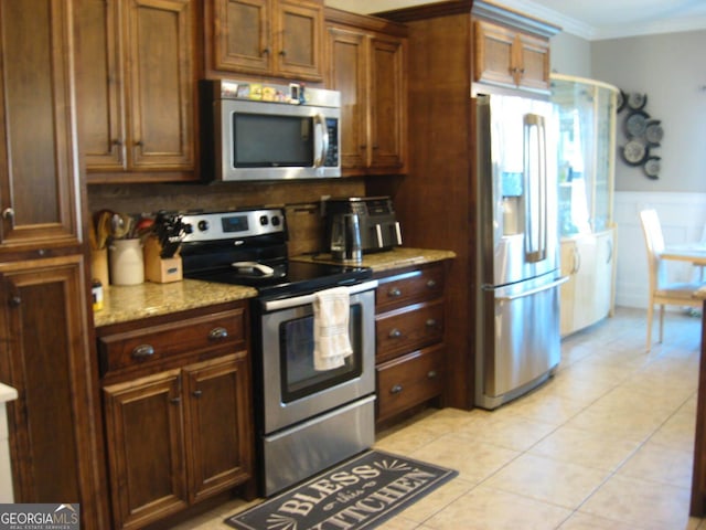 kitchen with light tile patterned floors, a wainscoted wall, ornamental molding, light stone counters, and stainless steel appliances