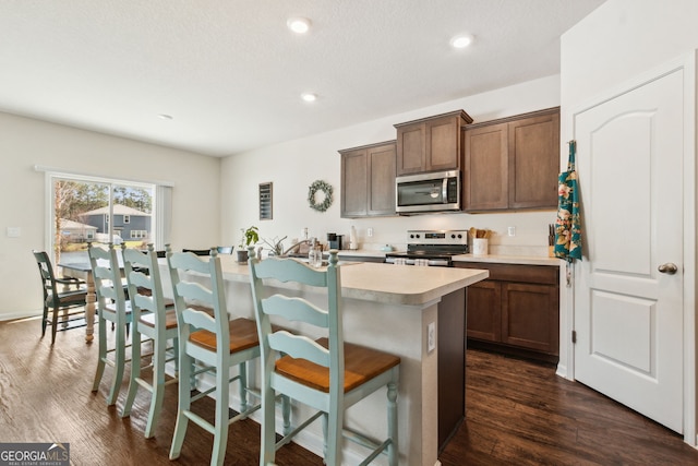 kitchen with stainless steel appliances, a kitchen breakfast bar, light countertops, dark wood-style floors, and an island with sink