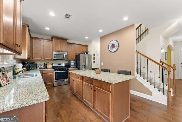 kitchen featuring stainless steel appliances, a kitchen island, a sink, brown cabinets, and dark wood-style floors