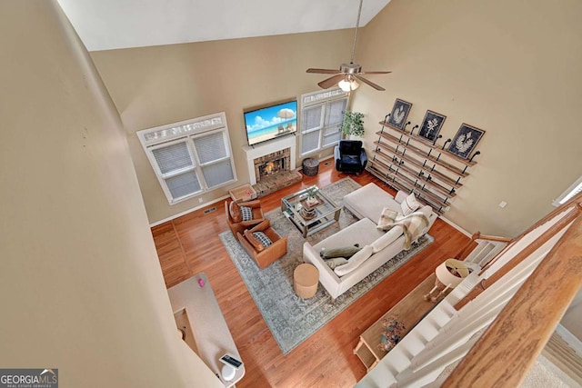 living area featuring ceiling fan, wood finished floors, high vaulted ceiling, a warm lit fireplace, and baseboards