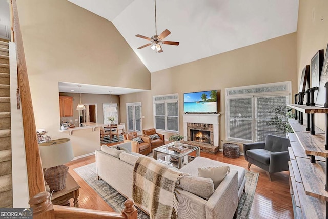 living area with high vaulted ceiling, a fireplace, a ceiling fan, light wood-style floors, and stairs