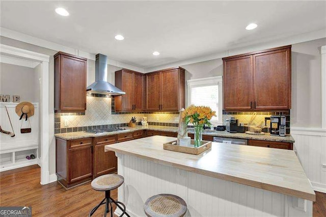 kitchen with gas stovetop, decorative backsplash, wall chimney range hood, wood finished floors, and a kitchen breakfast bar