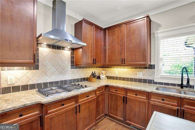 kitchen featuring tasteful backsplash, wall chimney exhaust hood, light stone counters, stainless steel gas cooktop, and a sink