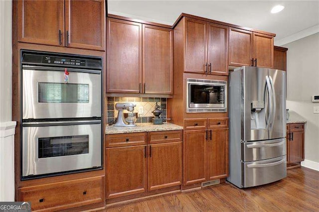 kitchen featuring tasteful backsplash, wood finished floors, appliances with stainless steel finishes, ornamental molding, and light stone countertops