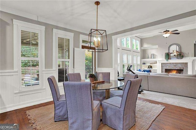 dining room with ceiling fan with notable chandelier, a lit fireplace, wood finished floors, and a decorative wall