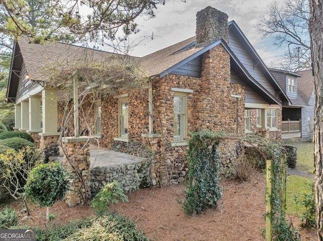 view of home's exterior with stone siding, a shingled roof, a chimney, and a patio