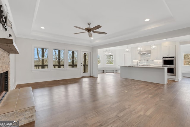 unfurnished living room with a large fireplace, baseboards, wood finished floors, a tray ceiling, and ceiling fan with notable chandelier