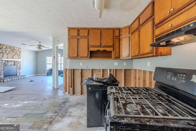 kitchen with a stone fireplace, black gas range, under cabinet range hood, a ceiling fan, and brown cabinetry