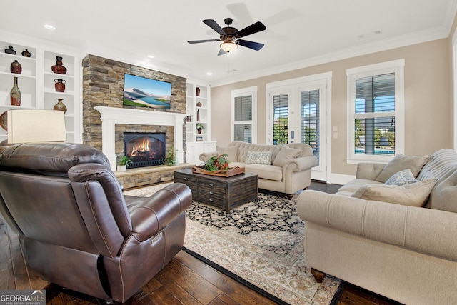 living area featuring built in features, a ceiling fan, dark wood-type flooring, crown molding, and a stone fireplace