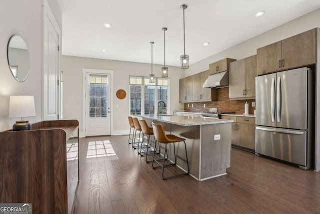 kitchen with under cabinet range hood, stainless steel appliances, dark wood-style flooring, a sink, and backsplash