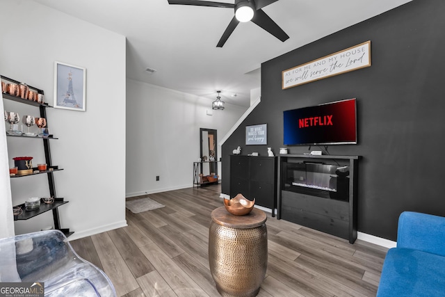 living area featuring ceiling fan, a glass covered fireplace, wood finished floors, and baseboards