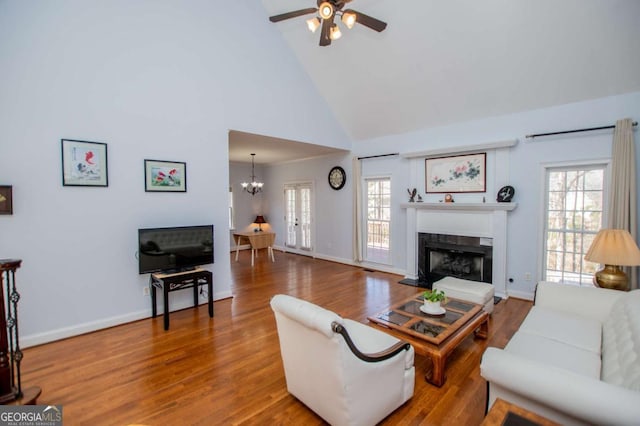 living area featuring a fireplace with raised hearth, wood finished floors, high vaulted ceiling, baseboards, and ceiling fan with notable chandelier