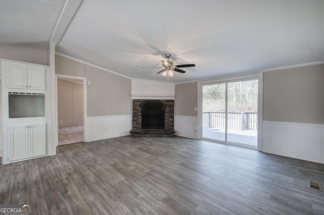 unfurnished living room with a wainscoted wall, ornamental molding, a stone fireplace, wood finished floors, and a ceiling fan