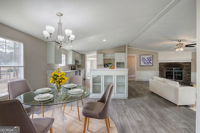 dining space featuring lofted ceiling, a stone fireplace, ceiling fan with notable chandelier, and light wood finished floors