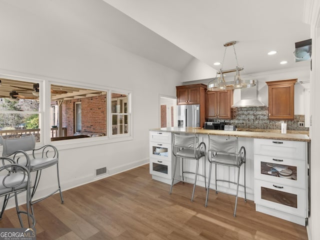 kitchen featuring brown cabinets, stainless steel refrigerator with ice dispenser, decorative backsplash, wall chimney range hood, and a kitchen bar