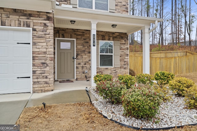 view of exterior entry with stone siding, covered porch, fence, and an attached garage